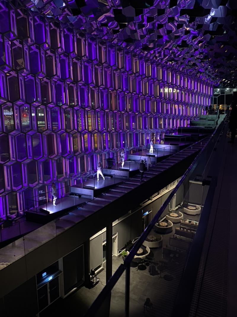 Dancers wearing white performing in front of large wall of purple lit windows