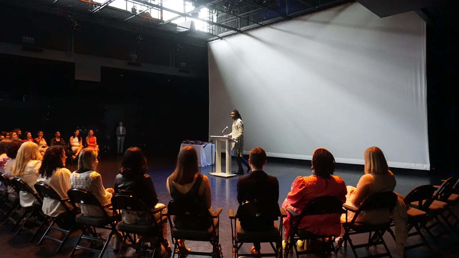 Students sitting on a stage listening to a speaker standing at a podium