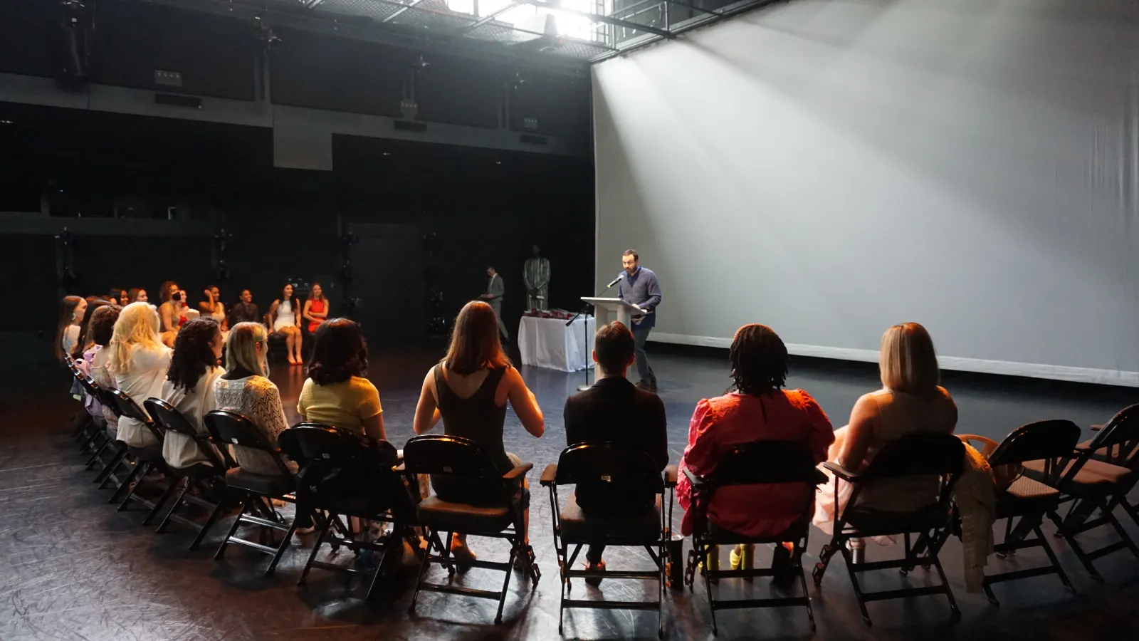 Students sitting on a stage listening to a speaker standing at a podium