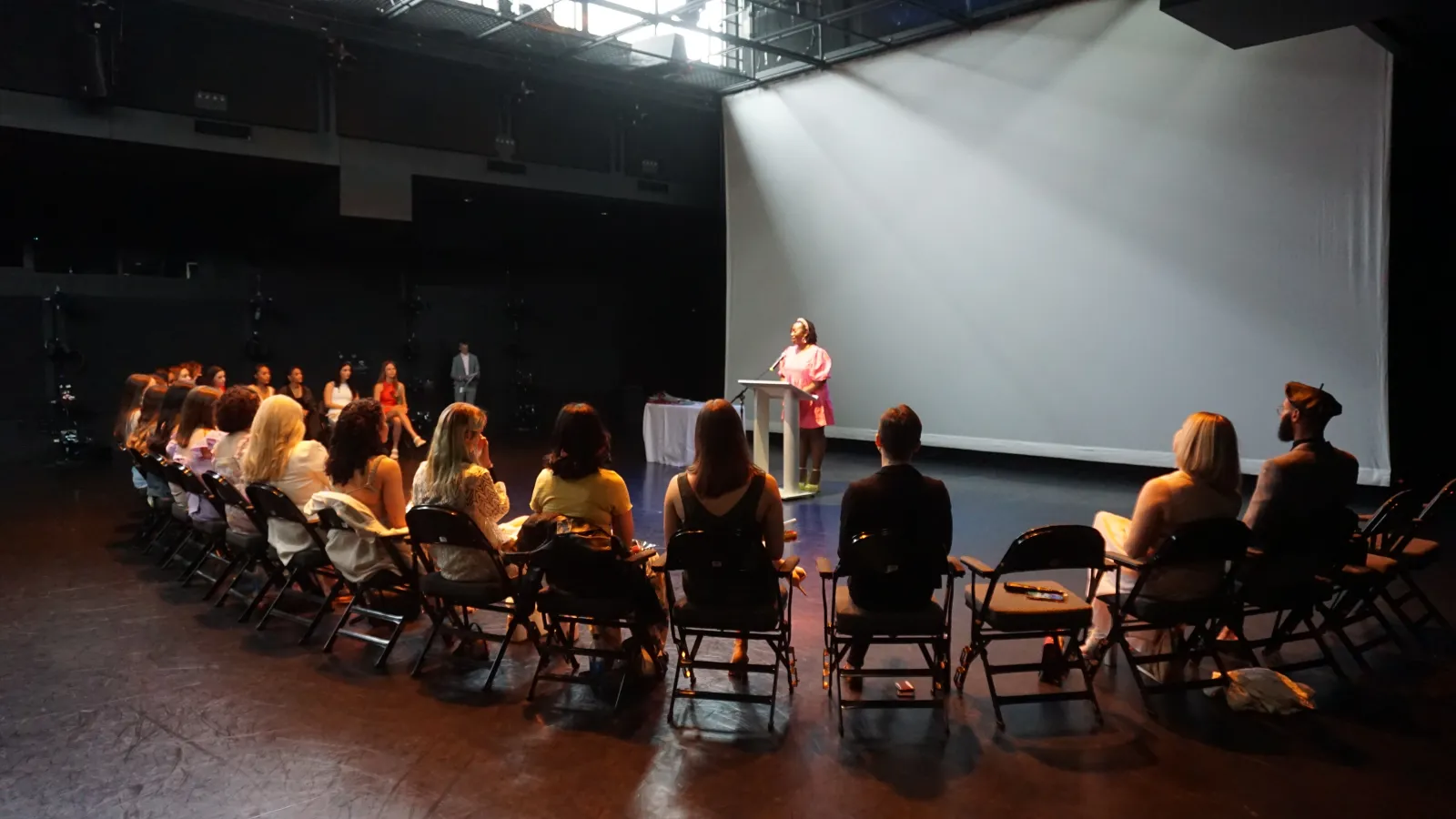 Students sitting on a stage listening to a speaker standing at a podium