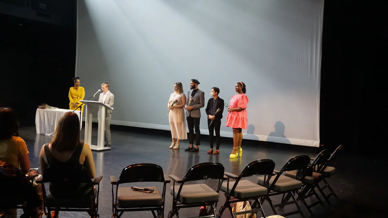 Students standing on a stage listening to a speaker standing at a podium