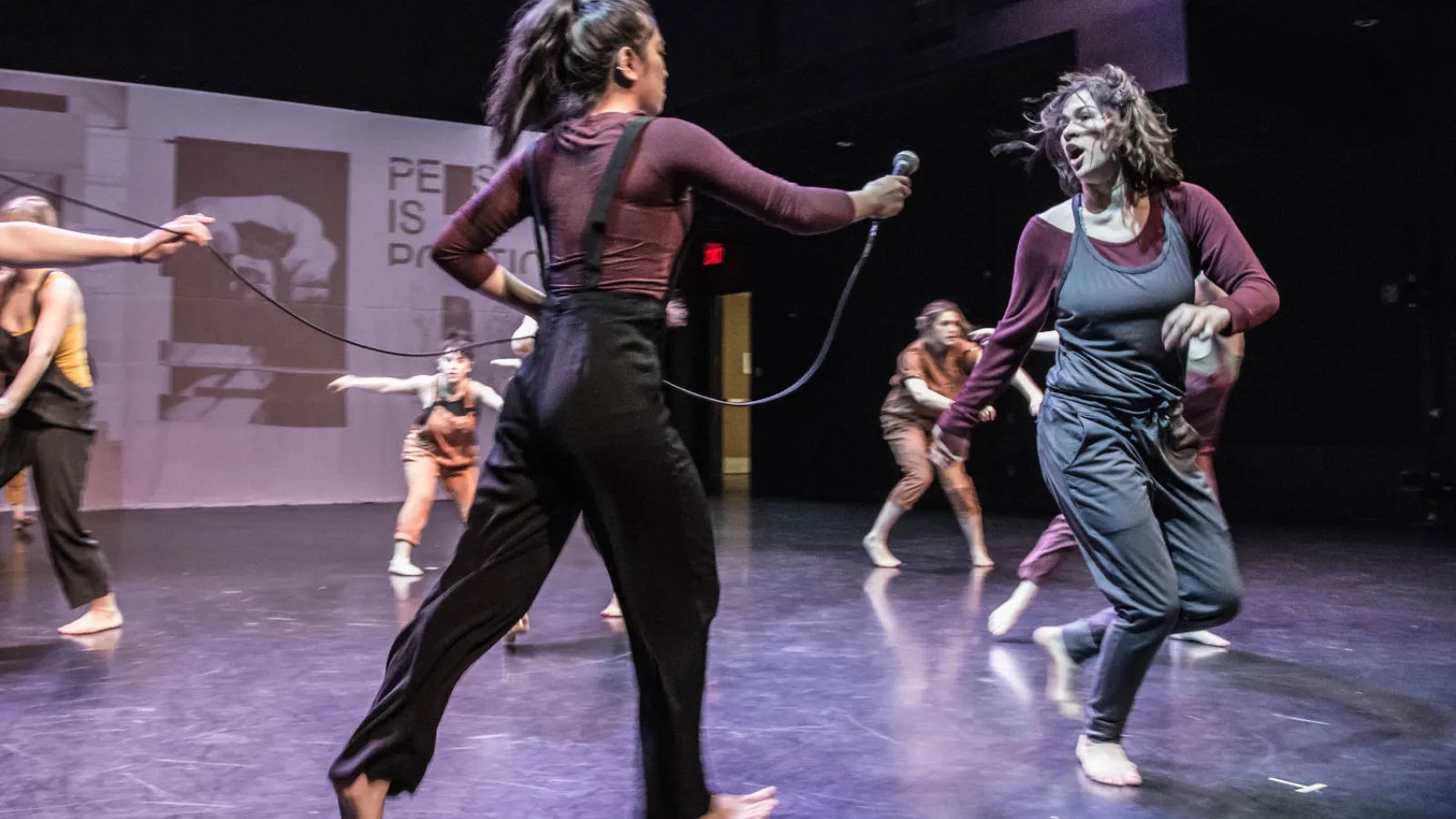 One dancer stretches iout her arms holding a microphone to another who is shouting and running backwards. Background performers contort as they move in front of a projection screen. 
