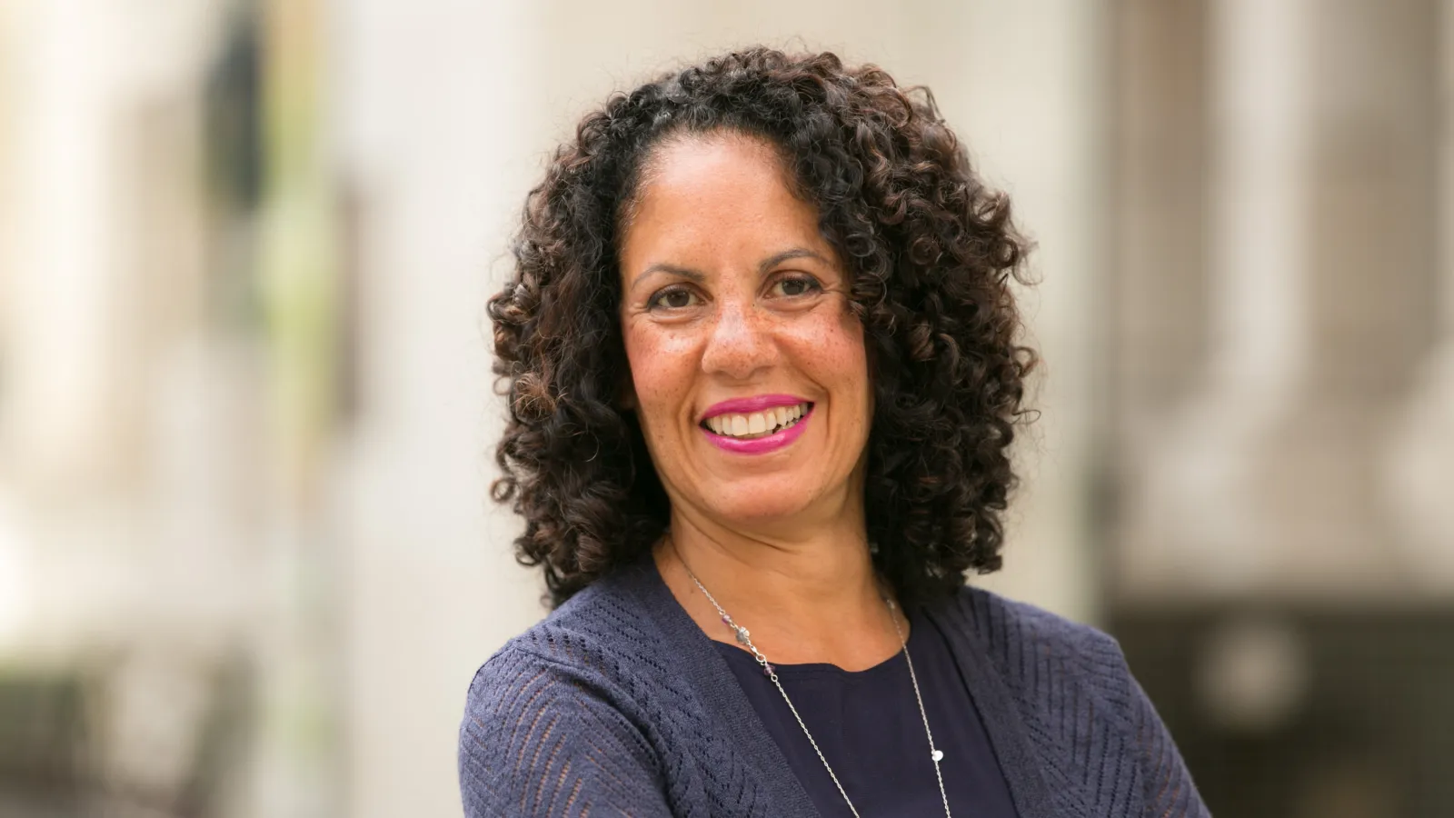 Woman with curly dark brown hair wearing blue shirt in front of blurred background