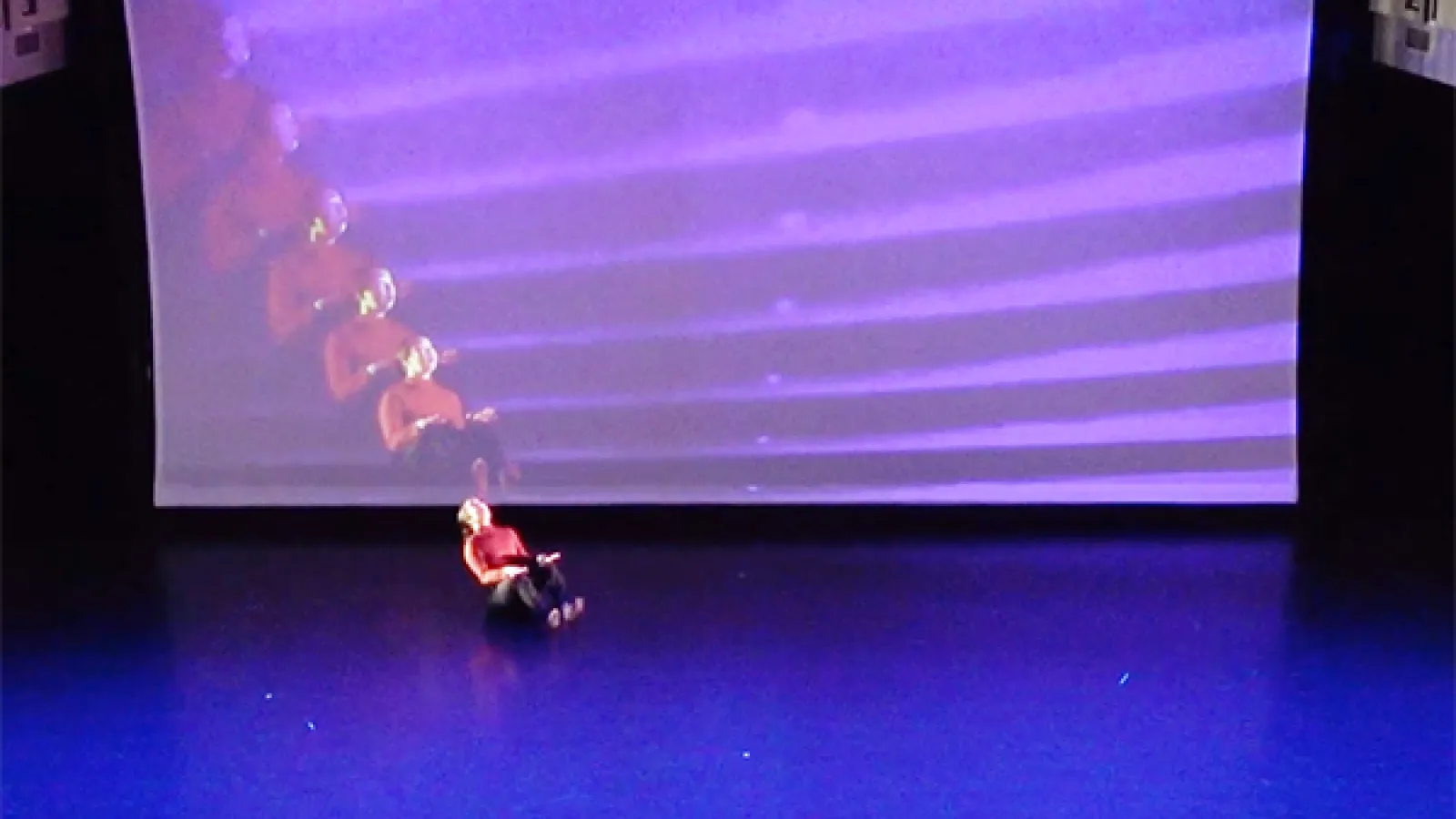 Dancer sitting on floor in front of screen with her image projected multiple times.