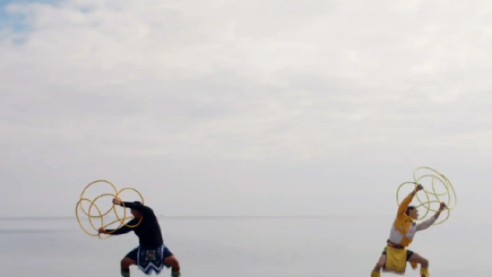 Dancers holding circular objects while performing on a beach with white clouds in the background