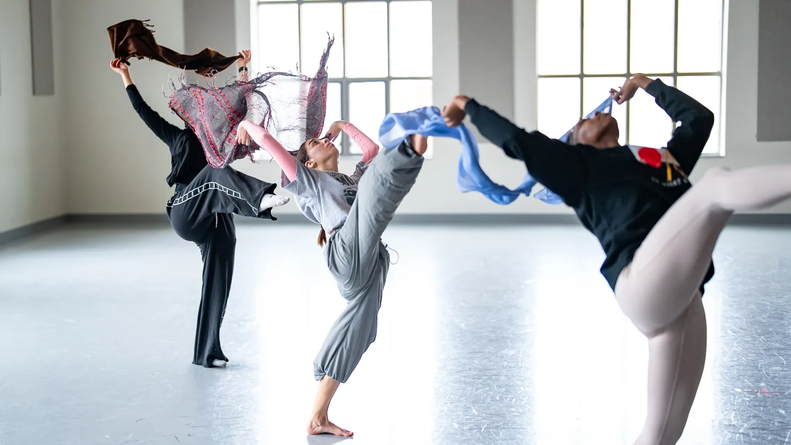 3 dancers leaning back with one leg extended waving scarves in a studio