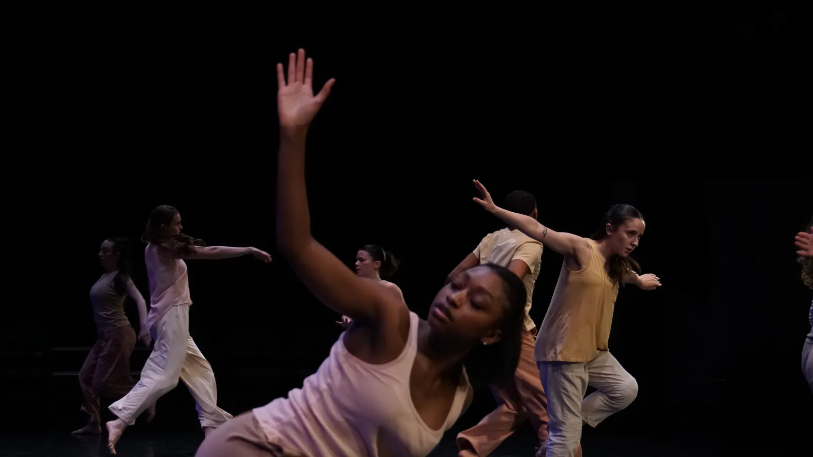 A dancer with dark skin and a light pink shirt reach towards the sky with an open palm as she leans over. In thr background many other dancers in similar colors are in motion. 
