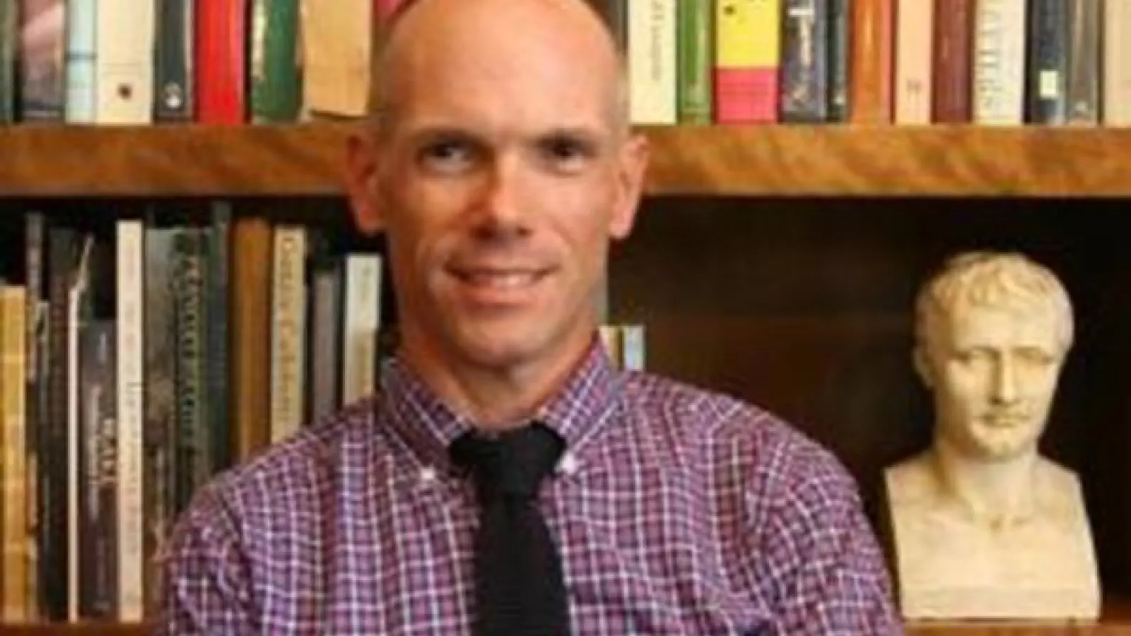 a bald person standing in front of shelves of books and a small bust statue
