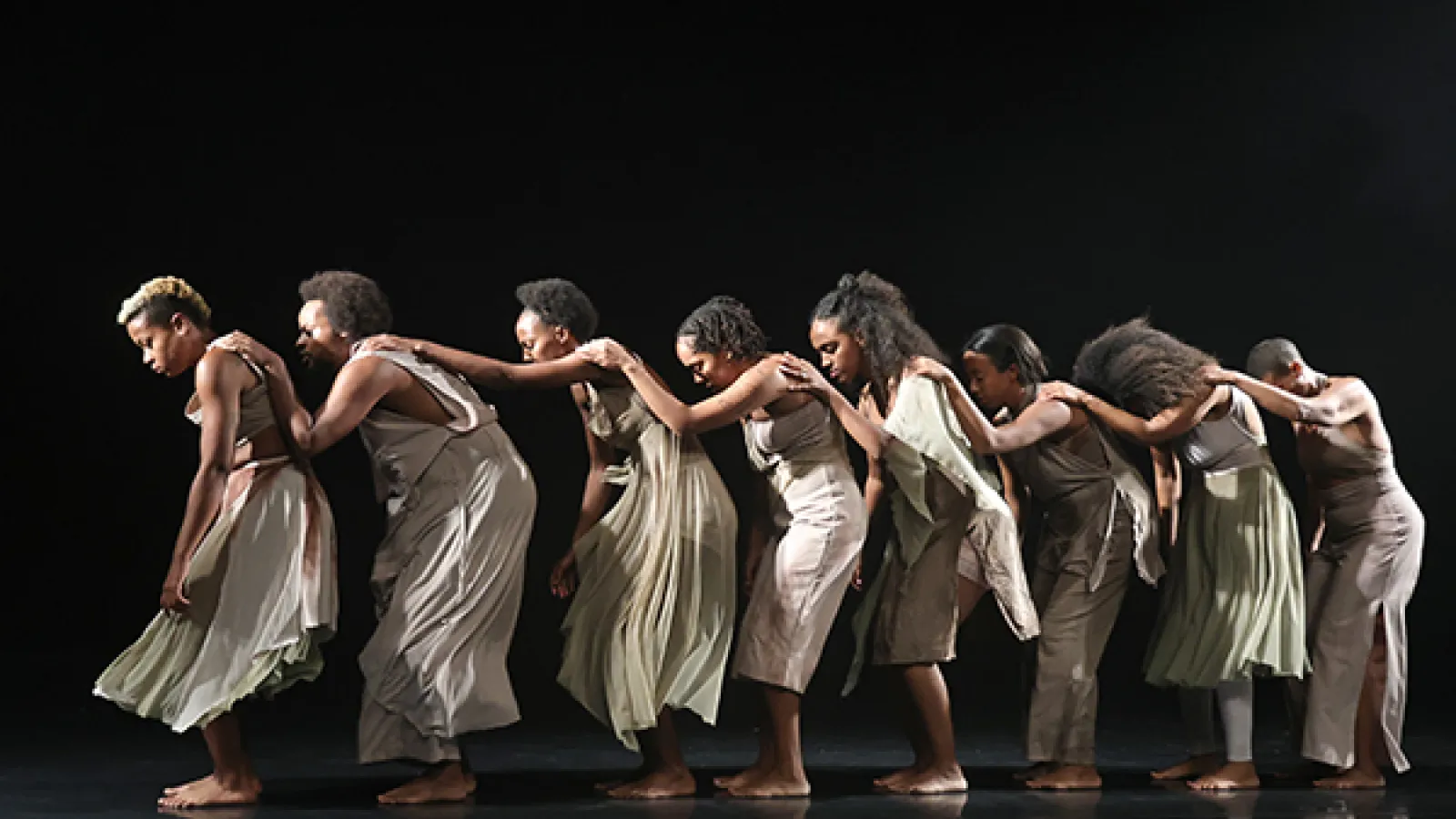 Eight women face to the left and lean slightly forward, each with their left hand on the shoulder of the person in front of them.
