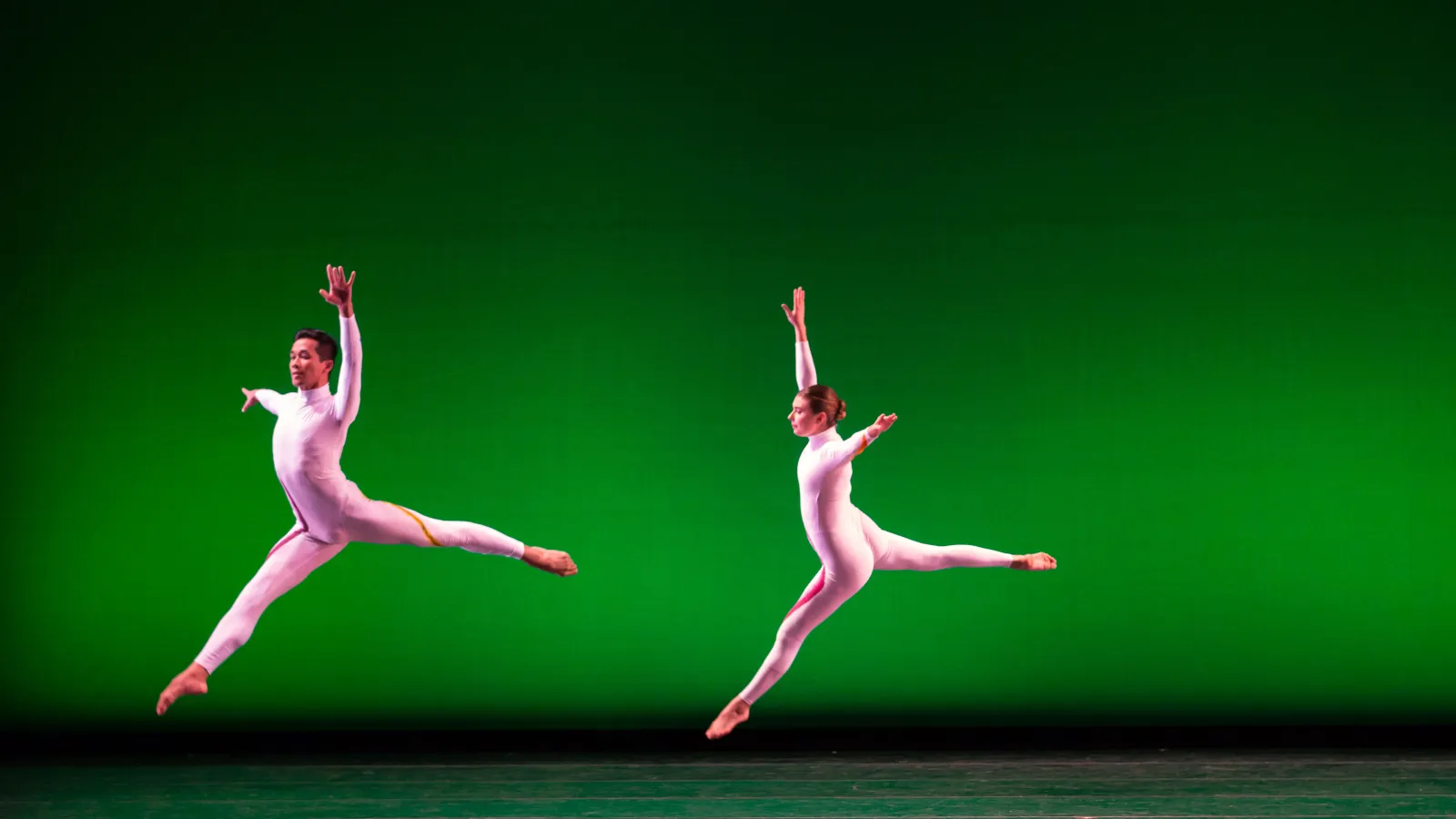 Two dancers in modern white costumes leaping in front of green background
