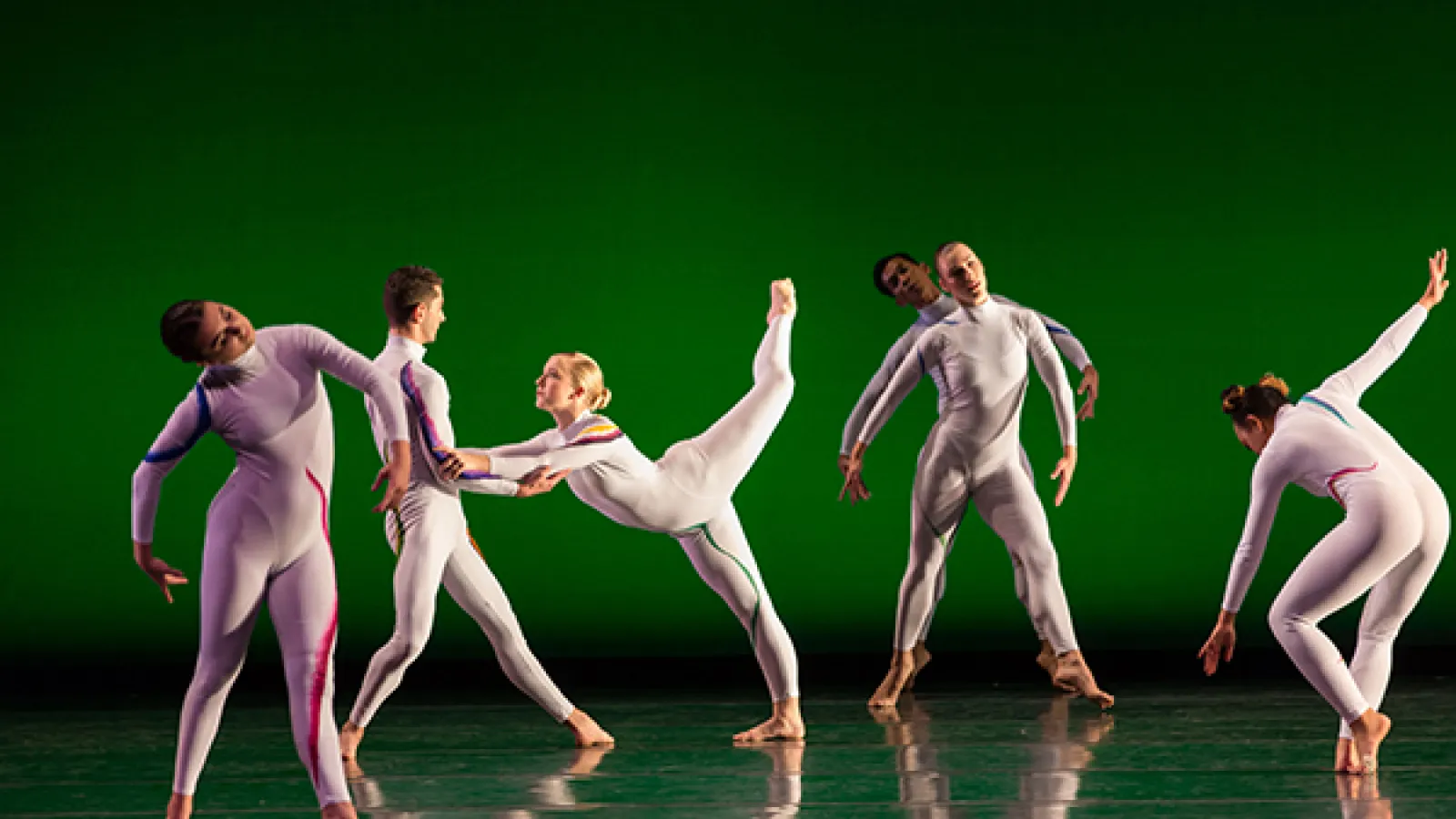 Six dancers in white modern costumes dancing on a stage with a green background