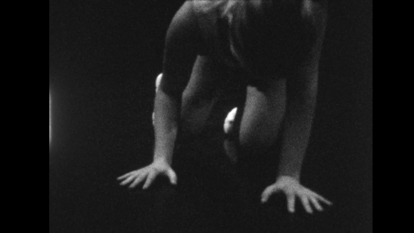 Black-and-white image shows a human figure bent forward, arms extended downward with hands splayed against a dark surface. The body is partially obscured, emphasizing tension in the limbs.