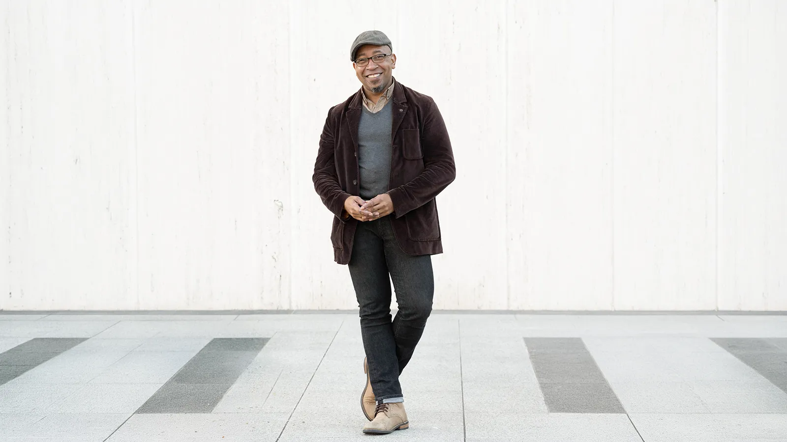 a man wearing a gray travelers cap, brown blazer, and blue jeans standing on a white wood floor with white wood paneling in the background