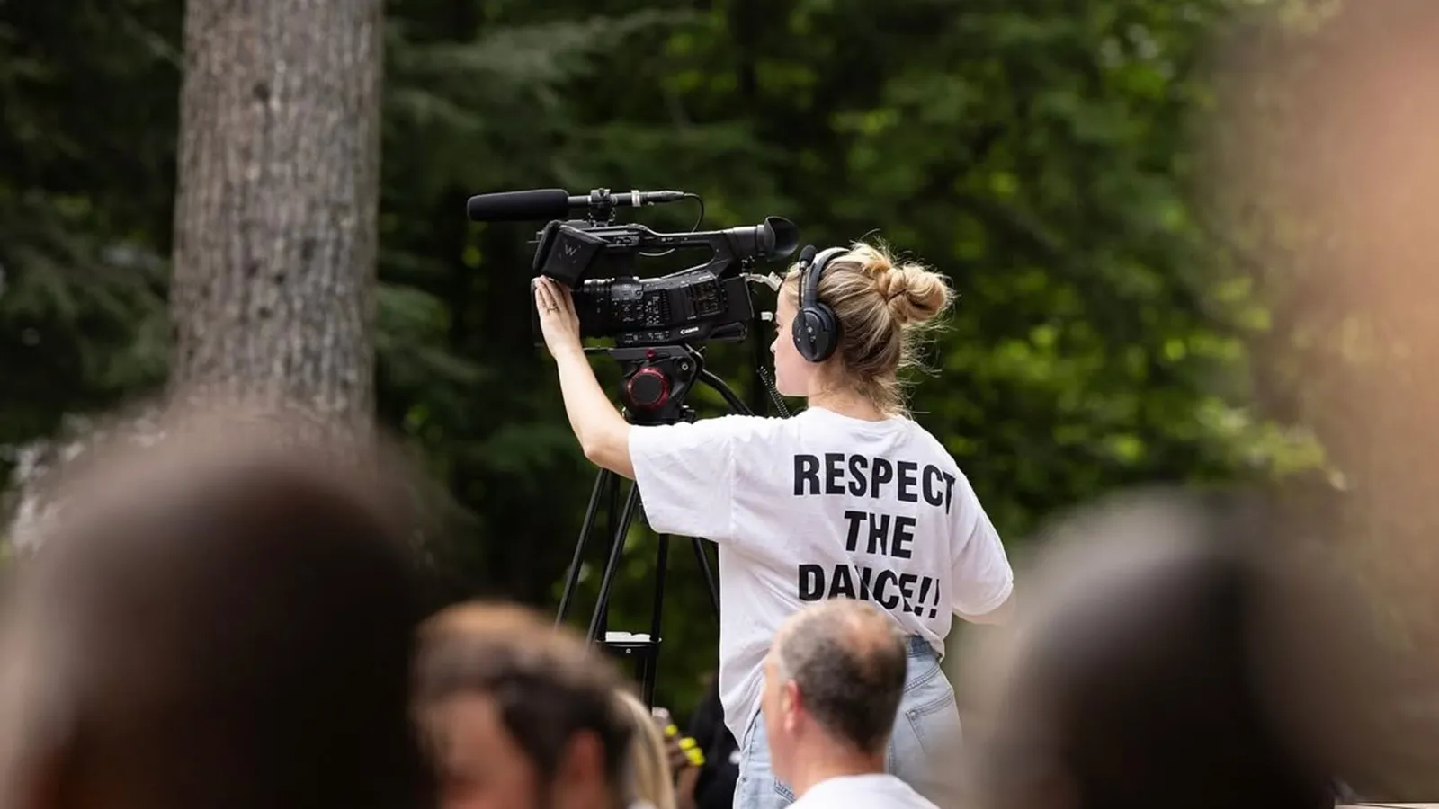 A woman operating a video camera outside with trees in the background
