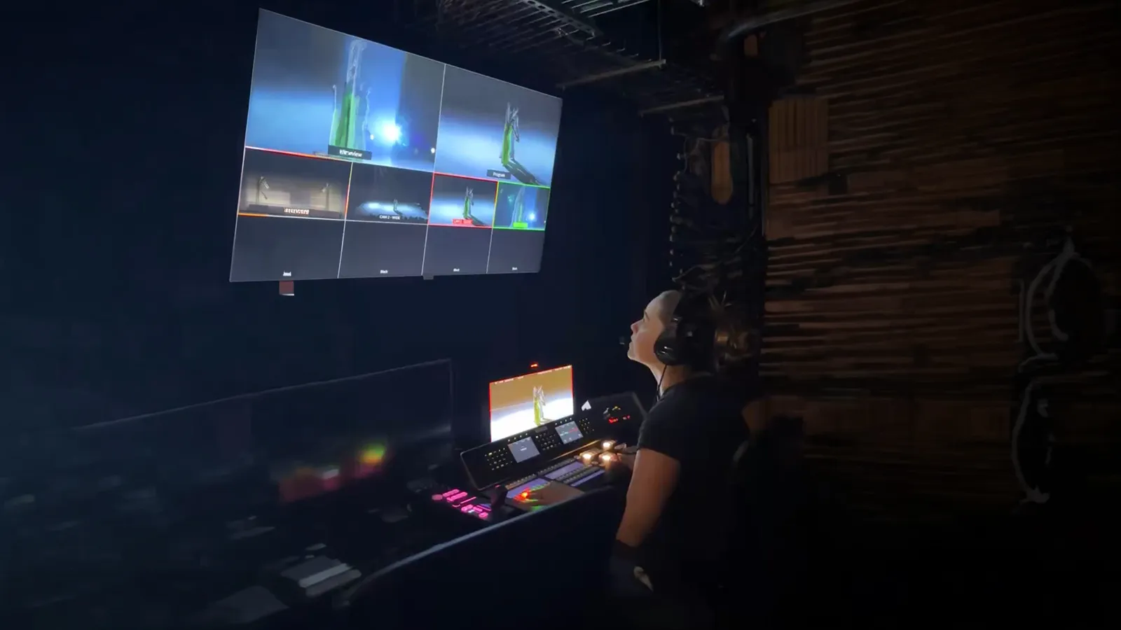 A woman in front in a dark theatre booth in front of a computer and tv monitors