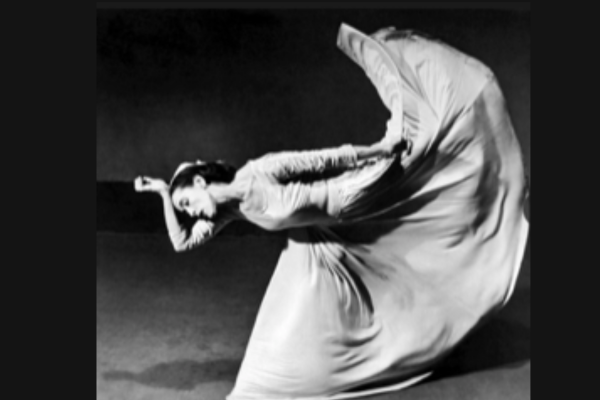 black and white photo of dancer in long white dress with one hand on her head and one leg lifted