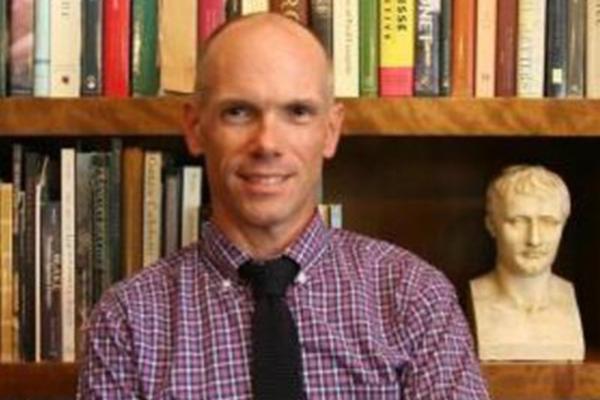 a bald person standing in front of shelves of books and a small bust statue