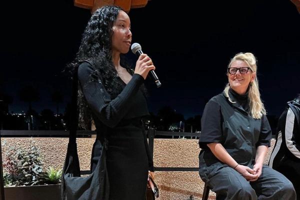 Person with long black hair and black dress standing with microphon next to person with long blonde hair sitting