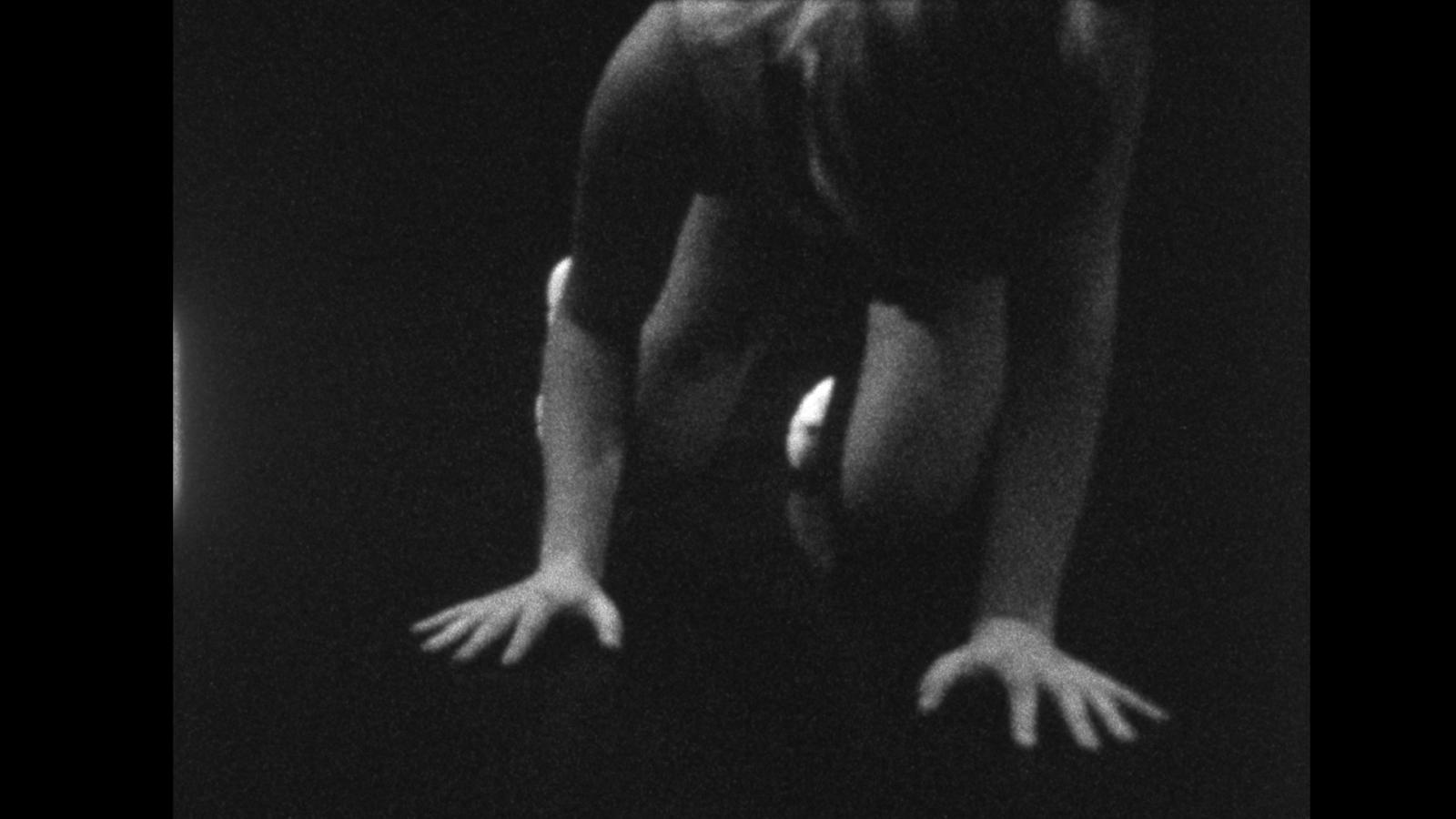 Black-and-white image shows a human figure bent forward, arms extended downward with hands splayed against a dark surface. The body is partially obscured, emphasizing tension in the limbs.