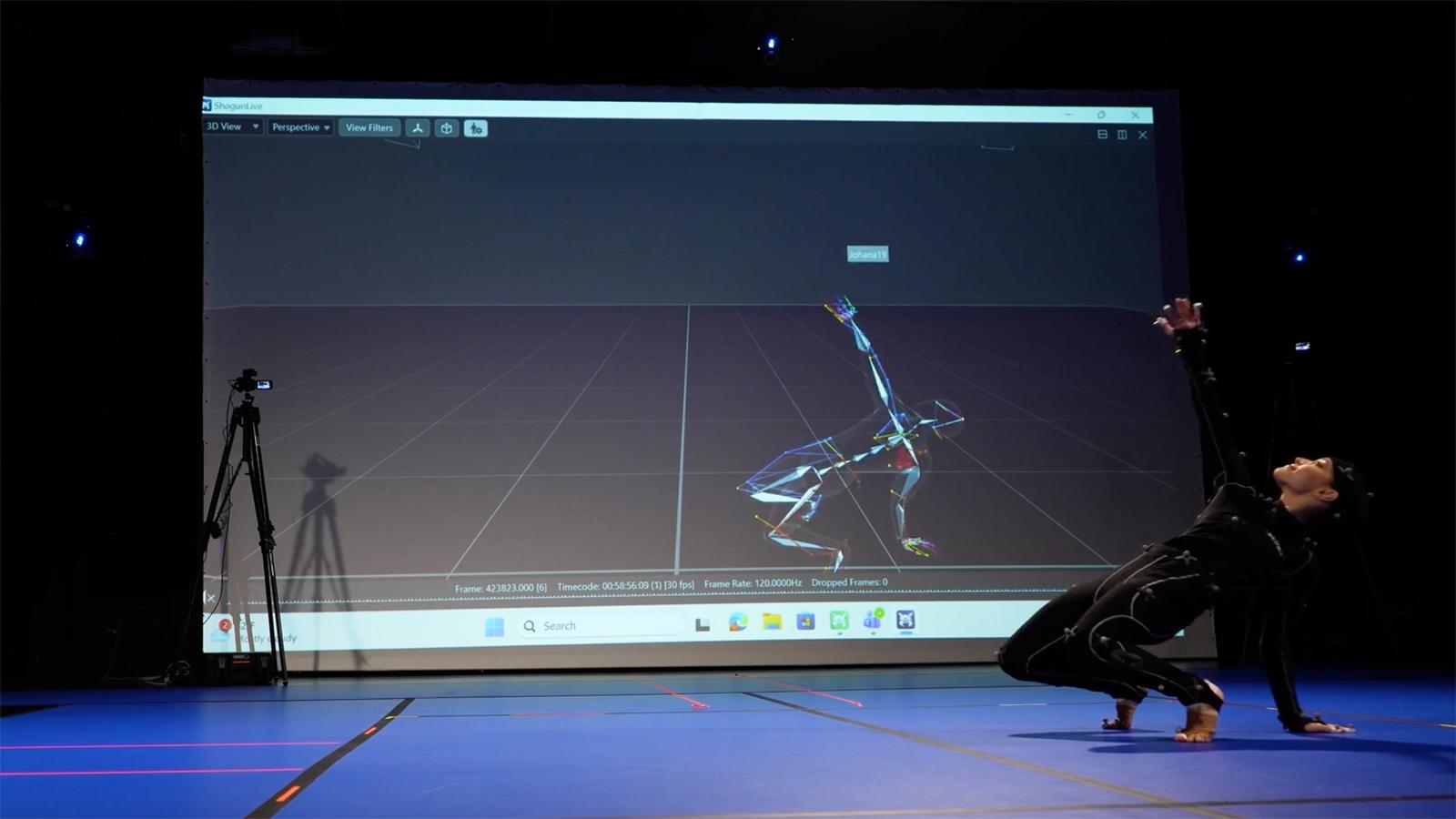 A dancer wearing a motion capture suit moving in front of a screen with her avatar projected on it