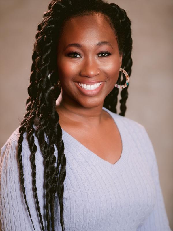 Kylee Smith, a black woman with long twists in her hair, resin hoop earrings, and a blue sweater looks into the camera and smiles fully showing her teeth.  