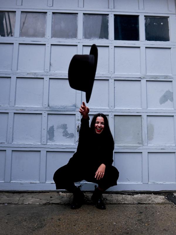exi squats in front of a blue door dressed in all black, she is smiling and tossing her hat at the camera. 