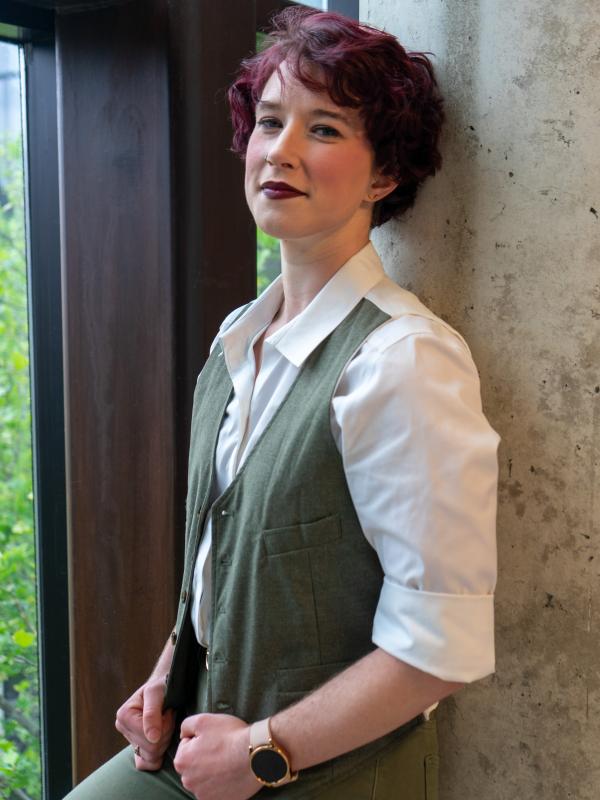 Person with purple hair and lipstick wearing a green vest leans against a concrete pillar and smiles at the camera.