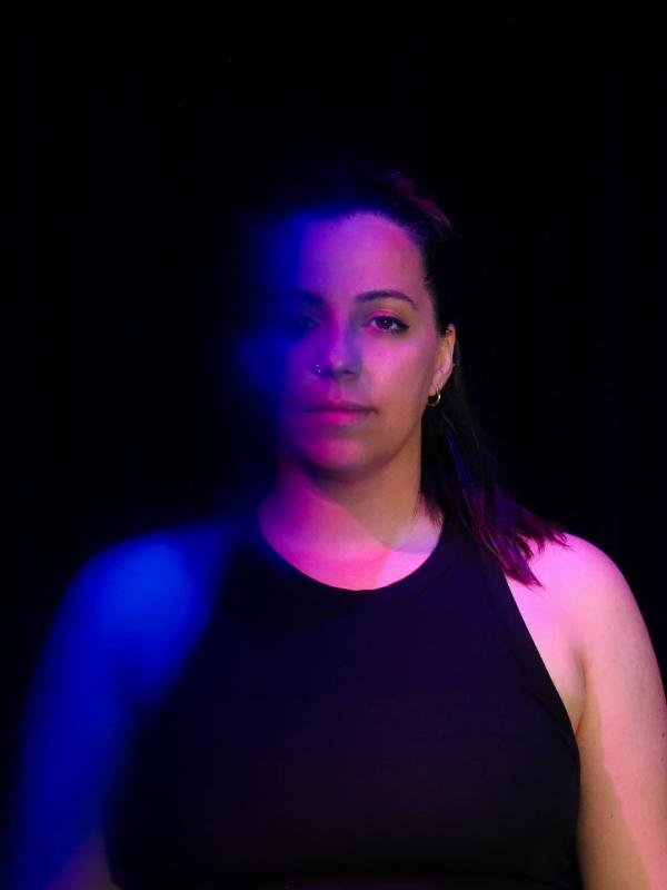 a white women with shoulder length brown hair looks at the camera with a blue and purple light cascading over half her face 