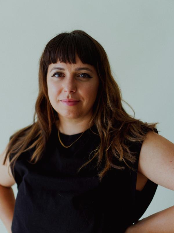 A white woman with shoulder length brown hair and bangs has her hands on her hips and smiles lightly at the camera 