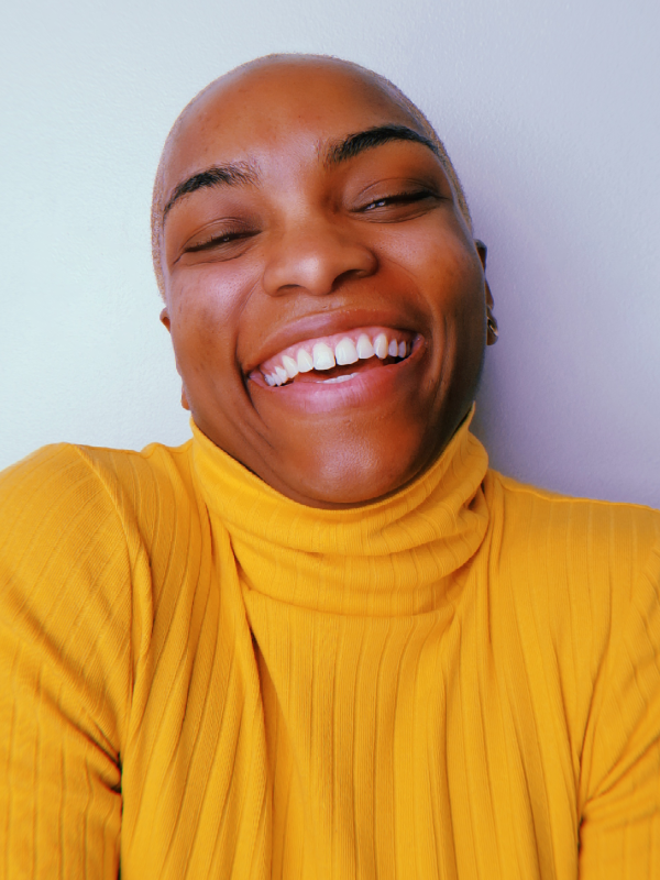 Portrait of a bald person with dark brown skin, wearing a mustard-yellow ribbed turtleneck, smiling widely and radiating happiness against light colored background
