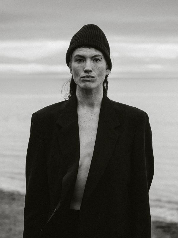 black and white photo of a woman in a black blazer and hat in front of the ocean 