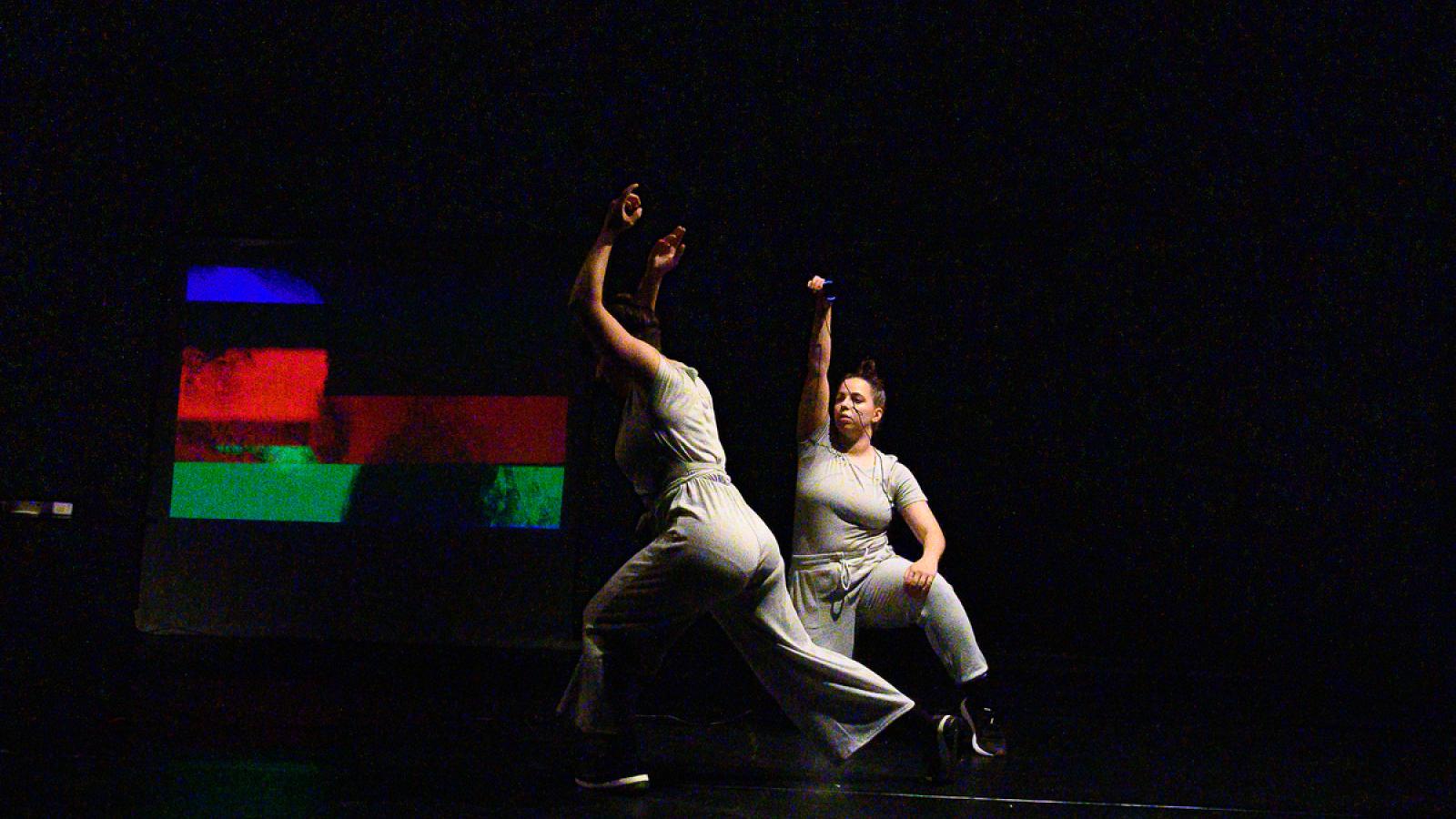  Two white dancers perform in front of a multicolored projection on stage dressed in gray clothing. One is holding a small camera above the other dancers head. 