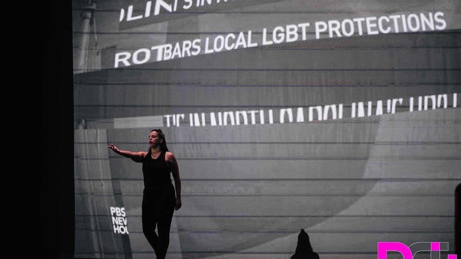  Lexi performs solo onstage in all back with her hand outstretched walking toewards the audience. A projection behind her shows  fractured words in black and white. 