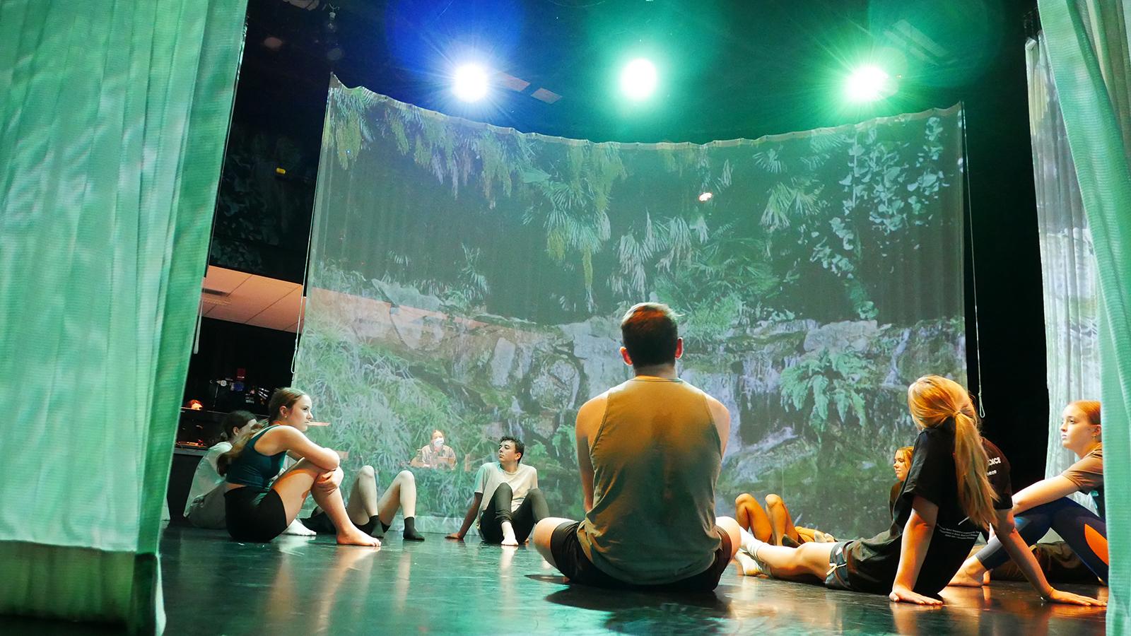 Dancers sitting on a studio floor talking with green scrims and stage lights