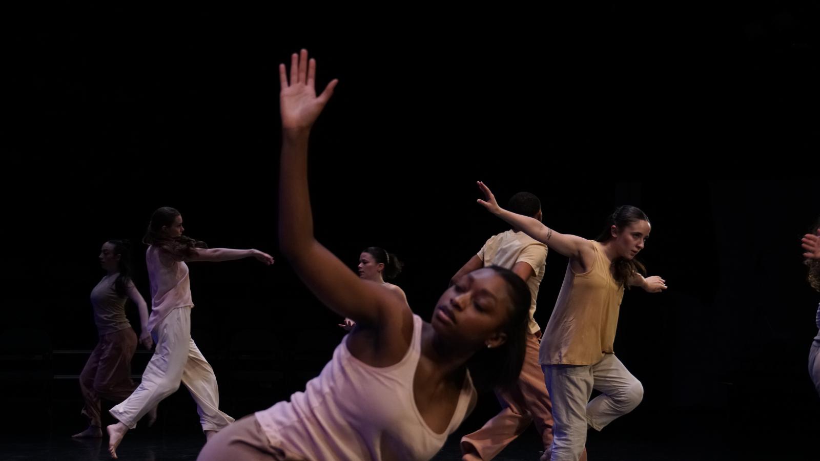 A dancer with dark skin and a light pink shirt reach towards the sky with an open palm as she leans over. In thr background many other dancers in similar colors are in motion. 