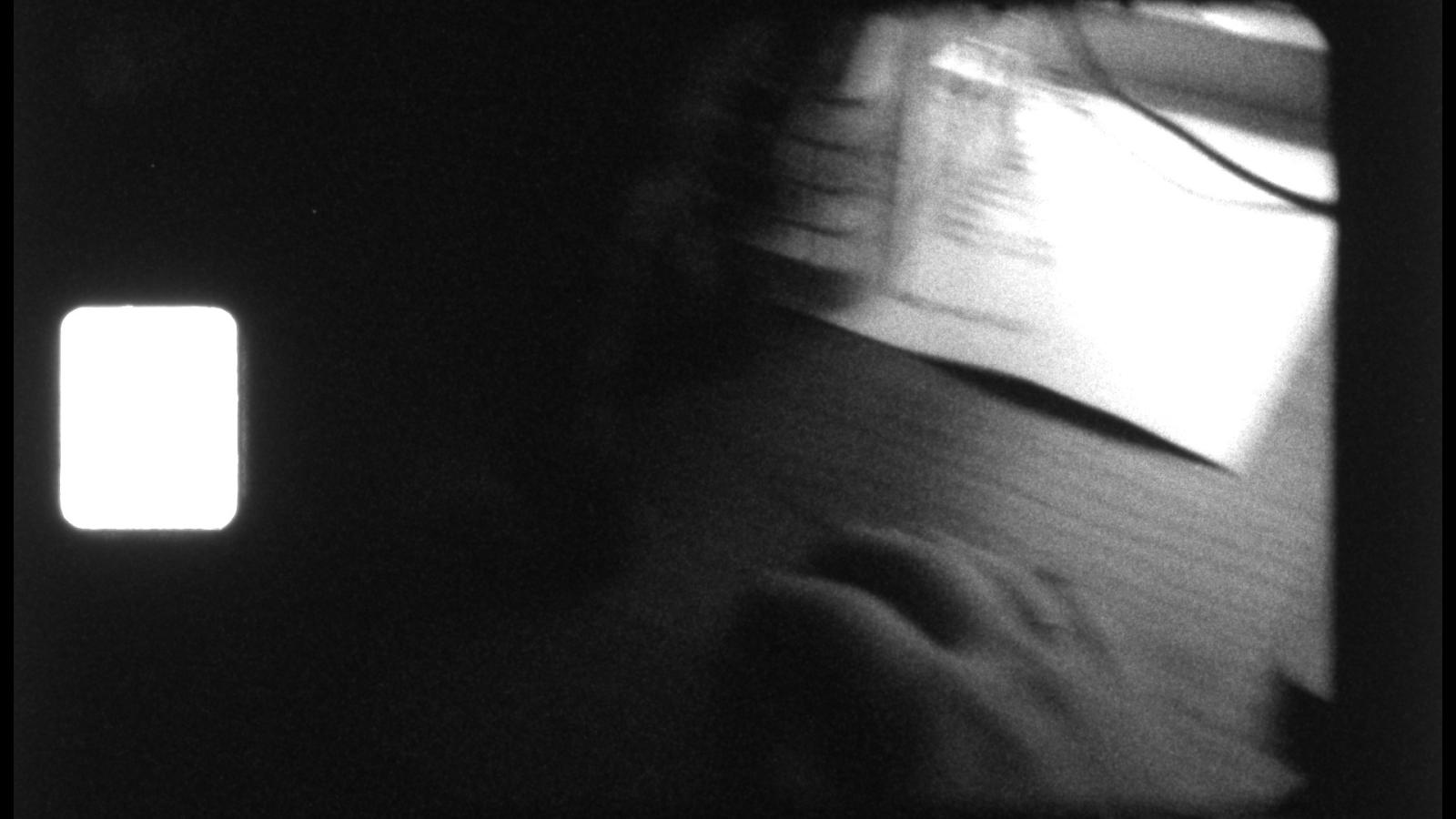 A black and white Super 8 film still of a hand in motion on a desk. Papers and cords are blurred behind her.