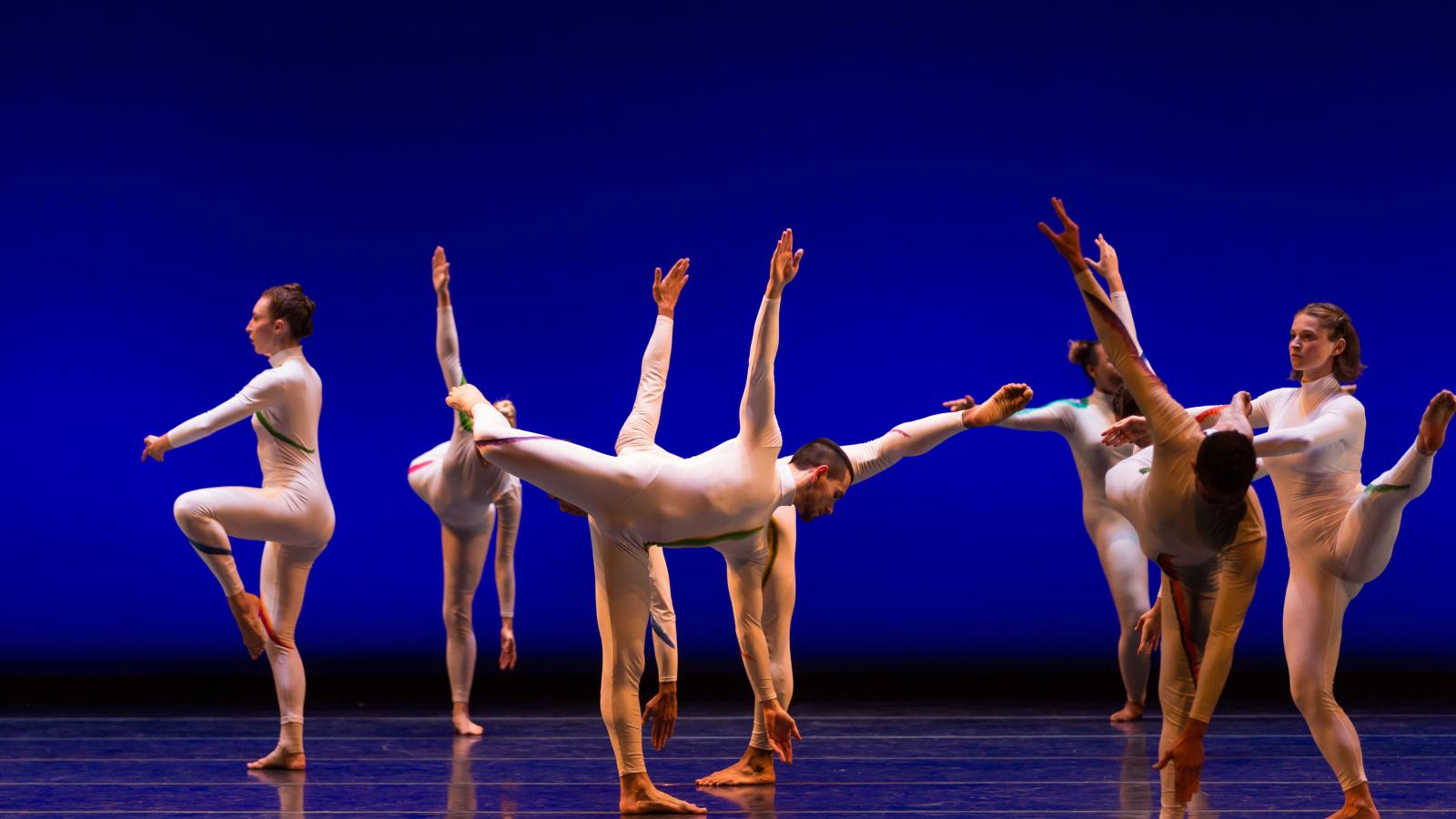 Seven dancers in modern white costumes dancing in front of blue background