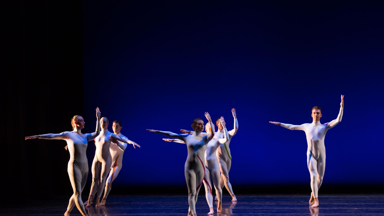 Seven dancers in modern white costumes dancing in front of blue background